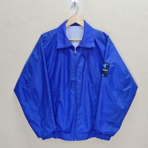 Puede incluir: Chaqueta azul brillante con cierre de cremallera y cuello. La chaqueta tiene un pequeño parche con el logotipo en blanco y negro en la manga izquierda. La chaqueta está colgada de una percha de madera.