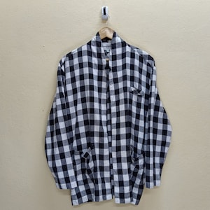 May include: A black and white checkered shirt hanging on a wooden hanger. The shirt has two front pockets and a collar. The pattern is a classic gingham design. The shirt is made of a lightweight fabric.