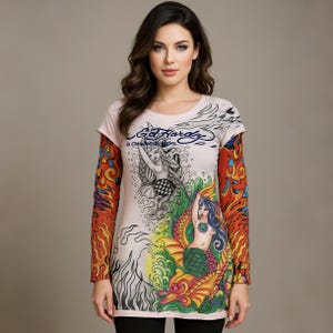 May include: A light pink long-sleeve shirt with a mermaid graphic and the text "Ed Hardy". The sleeves have a colorful flame design. The shirt features a crew neck and a relaxed fit.
