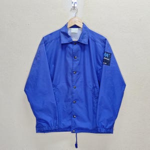 Puede incluir: Chaqueta azul brillante con botones y cuello. La chaqueta tiene un pequeño parche en la manga superior izquierda con el texto "HH Hansen A LIFE". La chaqueta está colgada de una percha de madera.