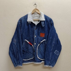 Puede incluir: Una chaqueta vaquera azul con forro de sherpa blanco. La chaqueta tiene cierre de botones, dos bolsillos de solapa y un parche en el pecho izquierdo que dice "Denim".