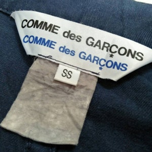 May include: A white tag with black text that reads "COMME des GARCONS* COMME des GARCONS*" and a smaller white tag with black text that reads "SS".