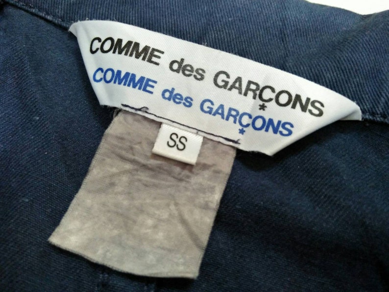 May include: A white tag with black text that reads "COMME des GARCONS* COMME des GARCONS*" and a smaller white tag with black text that reads "SS".