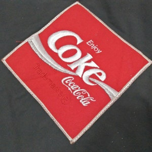 Może przedstawiać: Czerwony kwadratowy znaczek z białym haftowanym tekstem, kt&oacute;ry brzmi "Coke Coca-Cola" i "Enjoy" z symbolem zastrzeżonego znaku towarowego.