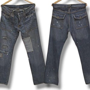 Puede incluir: Vaqueros de mezclilla azul oscuro con bolsillos delanteros y traseros. Los vaqueros tienen parches y salpicaduras de pintura. Los vaqueros cuelgan de una percha de madera.