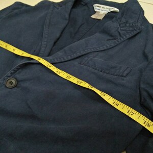May include: A dark blue blazer with a single button closure and a pocket on the left side. A yellow measuring tape is laid across the blazer, showing measurements in inches.