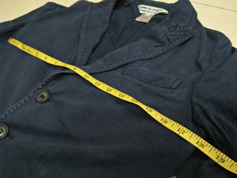 May include: A dark blue blazer with a single button closure and a pocket on the left side. A yellow measuring tape is laid across the blazer, showing measurements in inches.