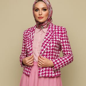 May include: A magenta and white houndstooth blazer. The blazer is open, revealing a light pink collared shirt and a matching pink skirt. A floral patterned headscarf completes the outfit. The background is a solid beige color.