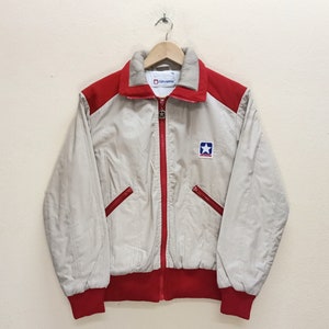 Puede incluir: Una chaqueta vintage beige y roja con cierre de cremallera y un logotipo de estrella azul que dice "Converse" en la parte delantera.