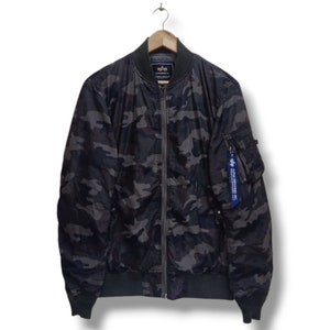 Könnte beinhalten: Eine schwarze und graue Camouflage-Bomberjacke mit Reißverschluss und einem blauen Etikett mit der Aufschrift "ALPHA INDUSTRIES INC."