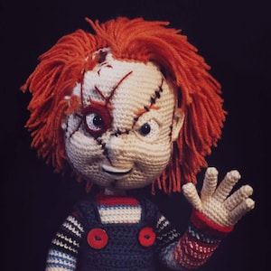 May include: A crocheted doll in the style of a horror character, with orange yarn hair and stitched details on the face. It wears blue dungarees with red buttons, striped sleeves, and red and white shoes. The background is black.