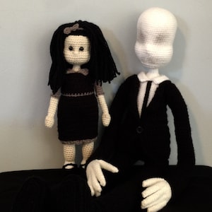 May include: Two handmade crochet dolls. One doll is a girl with black hair, a black dress, and gray accents. The other doll is a tall figure in a black suit with a white head and hands.