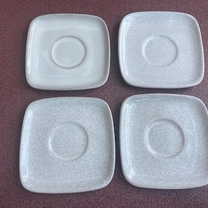 May include: Set of four white ceramic saucers with a speckled pattern. Each saucer has a small, round depression in the center.