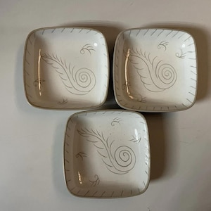 May include: Three square ceramic dishes with rounded corners, featuring an off-white speckled glaze and a hand-painted gray feather-like swirl design in the center. Each dish has a gray rim with short, evenly spaced vertical line accents.