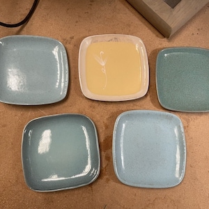 May include: Five square ceramic plates, four are light blue and one is white with a yellow center. The plates are arranged in a row on a brown surface.