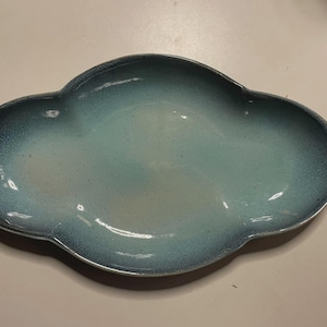 May include: An oval-shaped ceramic serving dish with a scalloped edge. The surface features a gradient glaze transitioning from a deep, speckled navy blue at the outer rim to a lighter, pale turquoise blue in the center with a smooth finish.