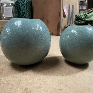 May include: Two teal ceramic vases, one larger than the other, with a speckled finish. The vases are round and have a smooth, glossy surface.