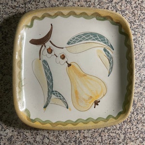 May include: A square ceramic plate with a yellow pear and green leaves painted on it. The plate has a scalloped edge and a yellow and green border.