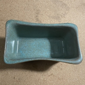 May include: A small, rectangular ceramic dish with a light blue glaze and a speckled finish. The dish is empty and has a slightly curved shape.
