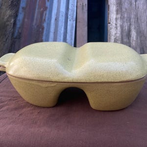 May include: A vintage, two-sectioned, yellow-green ceramic casserole dish with a speckled finish. The dish has a unique shape with rounded sides, a handle on each end, and a brown rim. It sits on a dark red-brown surface.