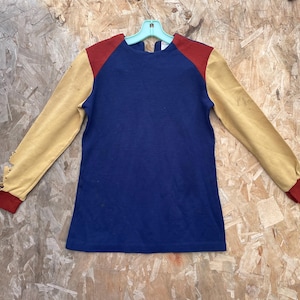 May include: A long-sleeved shirt with a color-block design. The shirt features a navy blue body, red shoulder accents, and tan sleeves with red cuffs. The shirt is hanging on a light blue hanger.