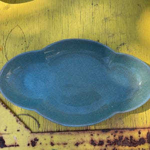 May include: A teal-colored, cloud-shaped ceramic dish. The dish has a textured surface and a slightly raised rim. It sits on a weathered yellow surface, creating a contrast in color and texture. The dish is likely used for serving food or as a decorative item.