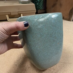 May include: A teal ceramic vase with a speckled glaze. The vase has a rounded, slightly asymmetrical shape.