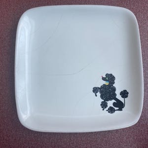 May include: White square plate with rounded corners. A black poodle illustration is on the right side of the plate. The poodle has a yellow collar and a pink nose. The plate has hairline cracks.