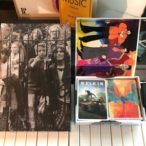 May include: A collection of vintage music-related items, including a black and white album cover featuring people behind a gate, a box with playing cards, and a colorful illustration of people. The items are placed on a piano.