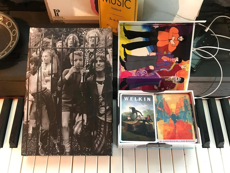 May include: A collection of vintage music-related items, including a black and white album cover featuring people behind a gate, a box with playing cards, and a colorful illustration of people. The items are placed on a piano.