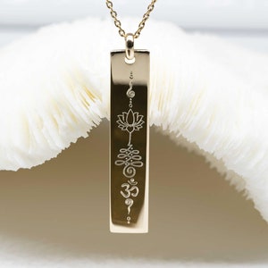 Könnte beinhalten: Goldkette mit einem vertikalen Barrenanhänger, auf dem eine Lotusblume, ein Knoten, eine Spirale und das Om-Symbol eingraviert sind.
