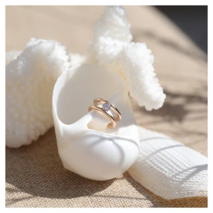 May include: A gold ring with a single clear stone set in the center. The ring is double-banded and sits on a white seashell.