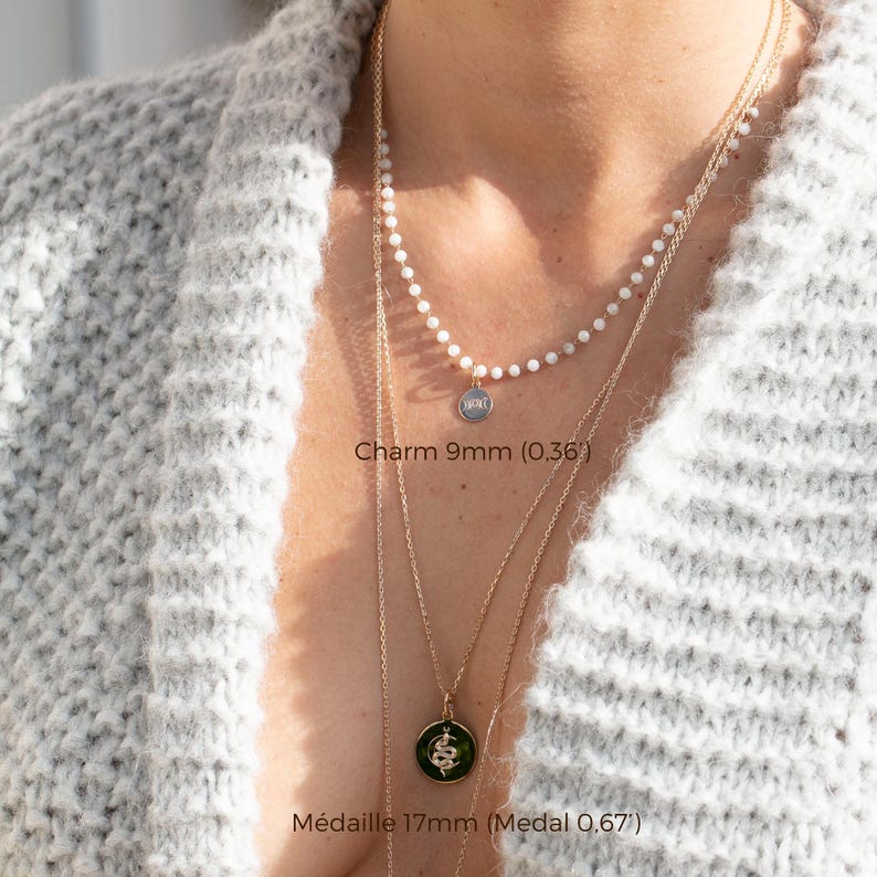 May include: A woman wearing a white sweater with two gold necklaces. The top necklace has a small silver charm with the letters "HOC" on it. The bottom necklace has a larger gold charm with a green snake design. The text "Charm 9mm (0.36)" is below the top charm and the text "M&eacute;daille 17mm (Medal 0.67)" is below the bottom charm.