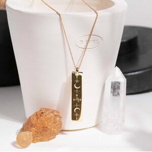 以下が含まれることがあります： 金色のペンダントネックレス。長方形のチャームには、天体のシンボルが描かれています。 ネックレスは白い鉢の前で、透明なクリスタルと琥珀色の石が近くにあります。 チャームには三日月、星、十字のシンボルがあります。
