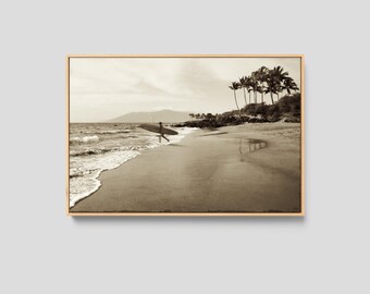 May include: A sepia-toned framed wall art print depicting a beach scene with a person carrying a surfboard toward the ocean. The landscape features a sandy shoreline, gentle waves, and a cluster of palm trees on the right side under a cloudy sky.