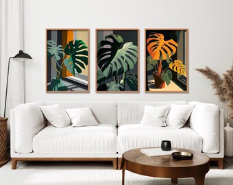 May include: A set of three rectangular wall art prints featuring stylized monstera plants in pots. The illustrations use a palette of green, orange, yellow, and dark gray tones, displayed in light brown wooden frames arranged horizontally on a wall.