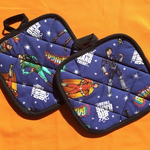 May include: Two blue quilted pot holders with black trim. The pot holders feature a repeating pattern of stars, the text "The Big Bang Theory", and characters from the show.