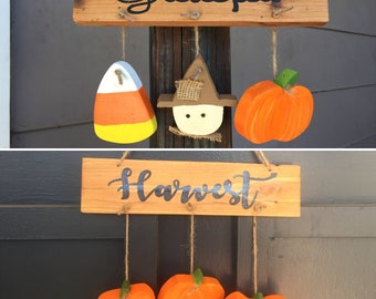 May include: A wooden sign with the word "Grateful" painted in black. Hanging from the sign are three decorations: a yellow and orange candy corn, a scarecrow with a brown hat, and an orange pumpkin. Below the sign is another wooden sign with the word "Harvest" painted in black. Hanging from this sign are four orange pumpkins.