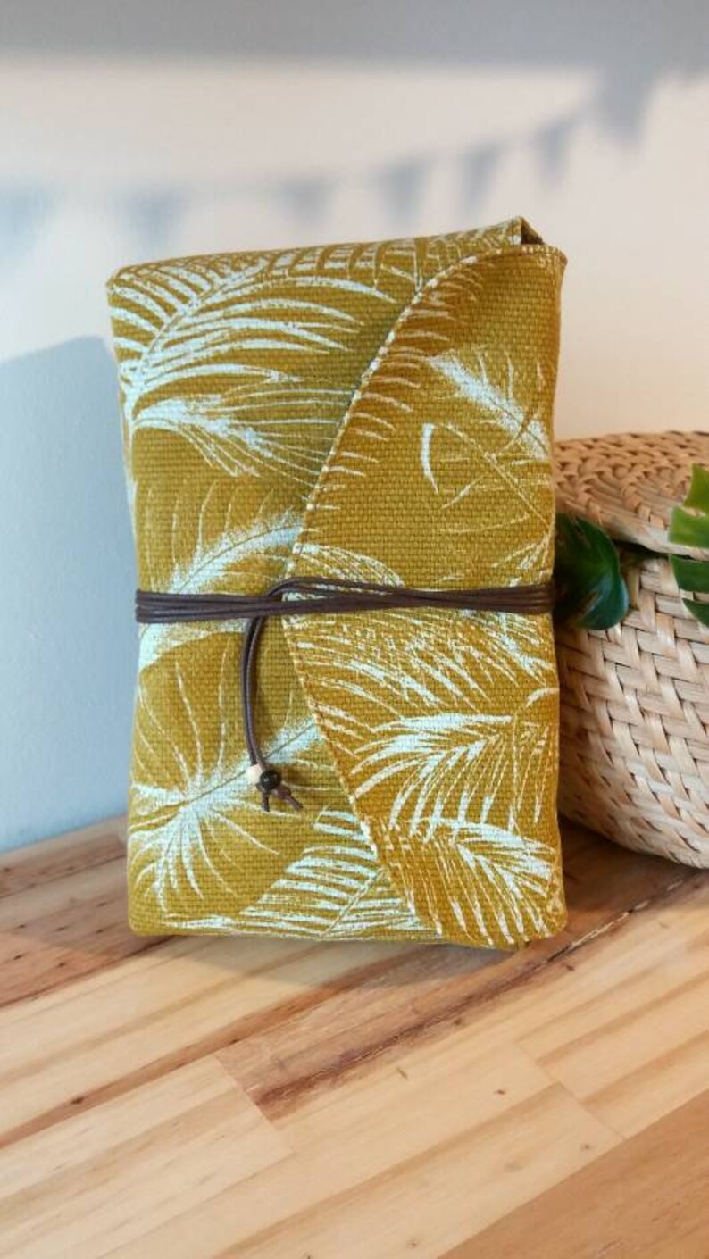 May include: A mustard yellow fabric pouch with a white tropical leaf pattern. The pouch has a brown leather strap and a white bead accent.