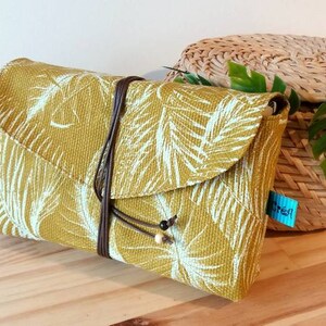 May include: A mustard yellow clutch with a white tropical leaf print and a brown leather strap. The clutch has a flap closure and is decorated with wooden beads.