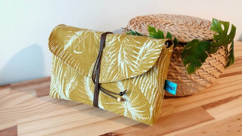 May include: A mustard yellow clutch with a white tropical leaf print and a brown leather strap. The clutch has a flap closure and is decorated with wooden beads.