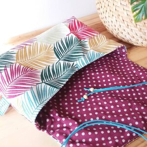 May include: A small, open, fabric pouch with a white background and a tropical leaf pattern in shades of green, pink, and yellow. The pouch is lined with a burgundy fabric with white polka dots. A turquoise cord is threaded through the pouch.