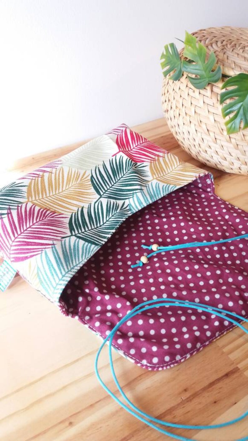 May include: A small, open, fabric pouch with a white background and a tropical leaf pattern in shades of green, pink, and yellow. The pouch is lined with a burgundy fabric with white polka dots. A turquoise cord is threaded through the pouch.