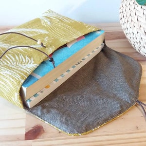 May include: A book sleeve made of green fabric with a white floral pattern. The sleeve has a brown drawstring closure and a brown lining. The sleeve is open and a book is inside.