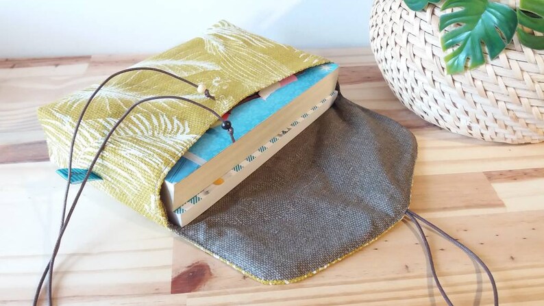 May include: A book sleeve made of green fabric with a white floral pattern. The sleeve has a brown drawstring closure and a brown lining. The sleeve is open and a book is inside.
