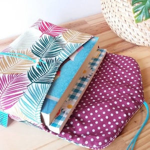 May include: A book sleeve with a tropical leaf pattern in shades of pink, green, and yellow. The sleeve is made of fabric and has a drawstring closure. The sleeve is open and a stack of books is inside. The sleeve is lined with a burgundy fabric with white polka dots.