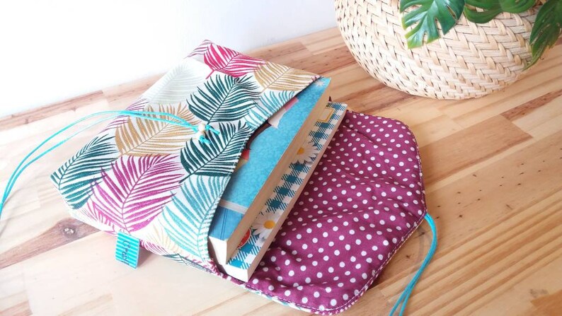 May include: A book sleeve with a tropical leaf pattern in shades of pink, green, and yellow. The sleeve is made of fabric and has a drawstring closure. The sleeve is open and a stack of books is inside. The sleeve is lined with a burgundy fabric with white polka dots.