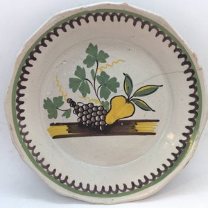 Peut inclure: Une assiette en céramique blanche avec un bord festonné et une bordure brune et verte. L'assiette présente un motif peint à la main d'une poire, de raisins et de feuilles dans des tons de jaune, vert et brun.