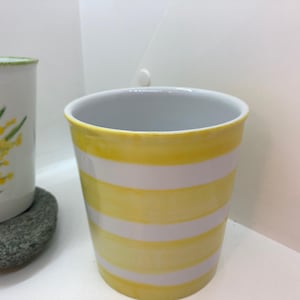 Könnte beinhalten: Zwei Keramikbecher, einer mit einem gelben und weißen Streifenmuster und der andere mit einem Blumenmuster mit gelben Blüten und grünen Blättern.