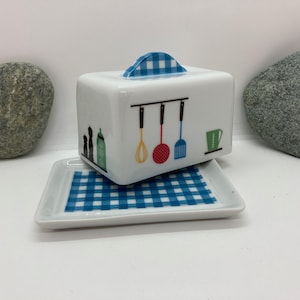 Può includere: Un porta burro in ceramica bianca con un coperchio a scacchi blu e bianchi. Il porta burro presenta un design di utensili da cucina, tra cui una frusta, una spatola e un mestolo rosso. Il porta burro è appoggiato su un vassoio a scacchi blu e bianchi abbinato.